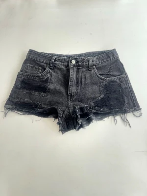 Svarta slitna jeansshorts - Säljer ett par svarta jeansshorts med råa kanter och slitningar framtill och baktill. Shortsen har fem fickor, bälteshällor och en knapp i midjan. Detalj med zebramönstrad patch bak. Perfekta för en edgy och avslappnad stil.