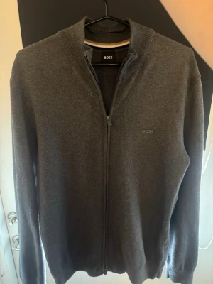 Hugo boss cardigan  - Snygg grå ziptröja från BOSS med diskret logga på bröstet. Tröjan har hög krage, hel dragkedja och ribbade muddar. Perfekt för lager-på-lager och enkel att matcha med andra plagg.