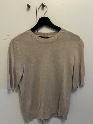 Beige kortärmad tröja från 157 - Stilren beige tröja från 157 med korta ärmar och rund halsringning. Tröjan är i ett mjukt, tunt stickat material och har ribbade muddar vid hals och nederkant. Perfekt för en avslappnad och trendig look.