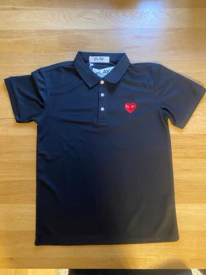 Svart pikétröja Comme des Garçons Play - Svart pikétröja från Comme des Garçons Play med klassisk krage, tre knappar och det ikoniska röda hjärtat broderat på bröstet. Perfekt för en stilren och trendig look.