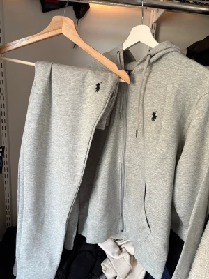 set från Polo Ralph Lauren - S  - Mysigt grått loungewear-set från Polo Ralph Lauren med zip-hoodie och matchande byxor. Setet har den klassiska broderade loggan på både tröjan och byxan. Tillverkat i mjukt bomullsmaterial, perfekt för chill dagar hemma men även på väg till gymmet.