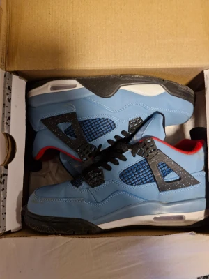 Jordan 4 Retro Travis Scott blå - Säljer ett par Jordan 4 Retro Travis Scott sneakers i blå mocka med svarta och röda detaljer. Skorna har svart nät på sidorna, svarta skosnören och vit/svart sula. Ikonisk Jumpman-logga på plösen och hängande tag. Perfekta för dig som gillar streetwear. 