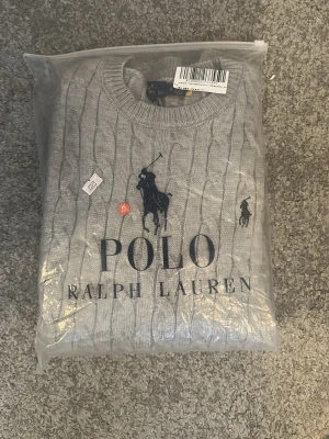 Grå kabelstickad tröja Polo Ralph Lauren - Kabelstickad grå tröja från Polo Ralph Lauren med rund halsringning och klassisk broderad logga på bröstet. Tröjan har en stilren look och är perfekt för lager-på-lager. Storlek M