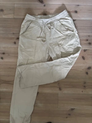 Beige mjukisbyxor från 157 - Beiga mjukisbyxor från 157 med resår i midjan och dragsko. Byxorna har två sidofickor och är tillverkade i ett mjukt och bekvämt material. Perfekta för en avslappnad stil och chill dagar.