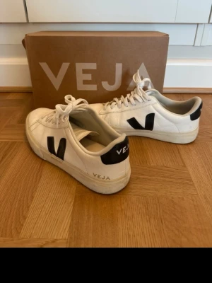 Vita Veja sneakers med svart detalj - Stilrena vita sneakers från Veja med svarta V-loggor på sidorna och svart detalj vid hälen. Skorna har klassisk låg profil, snörning och är tillverkade i läder med gummisula. Perfekta för en clean och modern look.