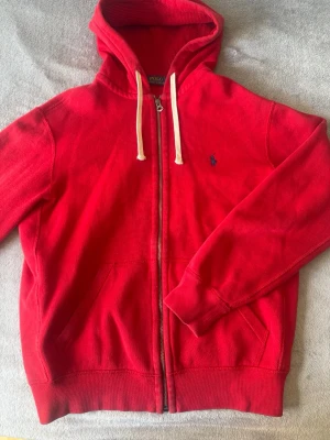 Röd hoodie från Polo Ralph Lauren - Snygg röd hoodie från Polo Ralph Lauren med dragkedja, vita snören i huvan och klassisk blå logga broderad på bröstet. Tillverkad i mjuk bomull och har två fickor framtill. Perfekt för en avslappnad och stilren look.