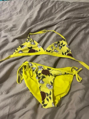 Gul blommig bikini med knyt - Snygg gul bikini med blommigt mönster i vitt och brunt. Överdelen har trekantsform och knyt i nacken och ryggen, medan underdelen har knyt i sidorna. Insidan av toppen är rosa. Perfekt inför sommaren🥰☀️
