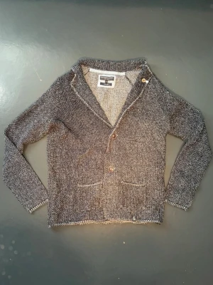 Stickad Kavaj - Mysig gråmelerad stickad kavaj med två knappar framtill och två framfickor. Kavajen har en avslappnad passform, sjalkrage och är tillverkad i grovstickat material. Perfekt för en chill och stilren look. Säljer då den är lite för liten för mig vid axlar och armar.