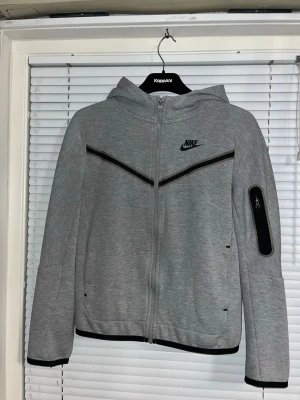 Grå Nike hoodie med dragkedja - Snygg grå hoodie från Nike med hel dragkedja och svarta detaljer. Har huva, två fickor fram och en extra ficka på armen. Tröjan är väldigt skön, men tyvärr så används den inte. Det är av den anledningen jag säljer denna tröja. Storleken är L i barn storlek. 
