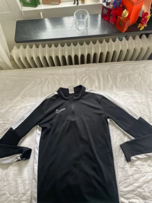 Svart Nike Dri-FIT träningströja - Säljer en svart långärmad träningströja från Nike med vita detaljer längs ärmarna och axlarna. Tröjan har halv dragkedja framtill och är tillverkad i Dri-FIT material som transporterar bort fukt. Perfekt för träning eller uppvärmning.