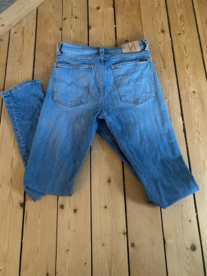 Blå jeans från Nudie Jeans - Säljer ett par klassiska blå jeans från Nudie Jeans med raka ben och femficksmodell. Jeansen har en ljus tvätt och subtila slitningar för en avslappnad look. Tillverkade i bomull med Nudie-logga på bakfickan.