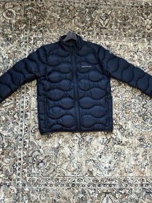 Mörkblå dunjacka från Peak Performance - Snygg mörkblå dunjacka från Peak Performance med vågformat quiltat mönster. Jackan har hög krage, dragkedja framtill och två sidofickor. Perfekt för kalla vinterdagar och riktigt lätt att bära. Klassisk och stilren design som funkar till det mesta.