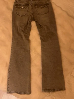 Grå bootcut jeans med fickdetaljer - Snygga grå bootcut jeans med klassiska fem fickor och dekorativa ficklock bak. Jeansen har en lätt utsvängd passform nedtill och är tillverkade i denim med en tvättad look. Perfekta för dig som gillar en avslappnad men trendig stil. Skriv om ni undrar nått!!💗💗