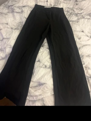 Svarta vida kostymbyxor - Säljer ett par svarta kostymbyxor med hög midja och vida ben. Byxorna har en klassisk look och är perfekta för en stilren outfit. Materialet känns mjukt och följsamt. Passar dig som gillar en avslappnad men ändå dressad stil. Behövs sys i bakre delen (se bild) 