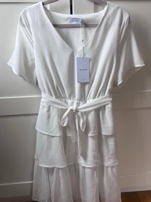 Vit omlottklänning från Vila - Säljer en vit omlottklänning från Vila med v-ringning, korta ärmar och volangdetaljer. Klänningen har ett knytband i midjan och är tillverkad i ett lätt, mjukt material som passar perfekt till sommaren.