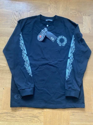 Svart långärmad topp från Chrome Hearts - Svart långärmad topp från Chrome Hearts med vita tryckdetaljer på bröstet och längs ärmarna. Rund halsringning och klassisk passform. Perfekt för dig som gillar streetwear och vill sticka ut med snygga detaljer.