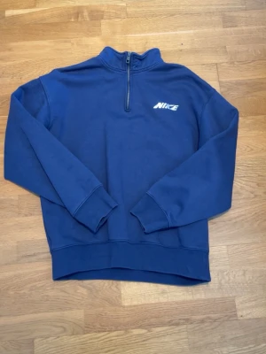 Blå Nike half-zip hoodie - En stilren blå hoodie från Nike med half-zip framtill och vit logga på bröstet. Tröjan har hög krage, långa ärmar och ribbade muddar. Perfekt för en avslappnad och sportig look. Man får en vintage känsla som passar perfekt vid alla årstider. Köpt för 1400 i butik men använder inte den längre, har däremot använt bara några fåtal gånger så den är så gott som ny. Den är också oversized när det gäller passform men kort i midjan så passar någon som vanligtvis har S/M.