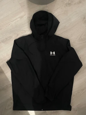 Under armour windbreaker - Snygg svart vindjacka från Under Armour med huva och dragsko. Jackan har dragkedja framtill och ett vitt logotryck på bröstet. Tillverkad i lätt och tåligt material med meshfoder i huvan för extra komfort. Perfekt för blåsiga dagar.