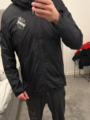 Svart AIK vindjacka från Nike - Svart vindjacka från Nike med AIK:s klubbmärke på bröstet och diskret Nike-logga. Jackan har huva, dragkedja framtill och är tillverkad i lätt, vindtätt material. Perfekt för dig som vill visa ditt AIK-stöd med stil.