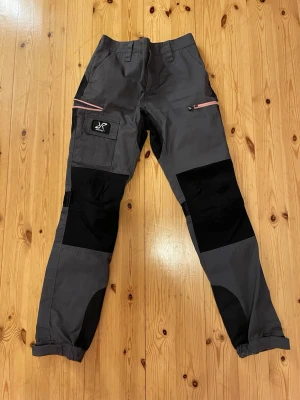 Grå fritidsbyxor från Revolution Race - Snygga grå fritidsbyxor från Revolution Race med svarta förstärkningar på knän och bak, samt rosa dragkedjor på fickorna. Byxorna har flera praktiska fickor och justerbara benslut med kardborre. Perfekta för en aktiv stil!