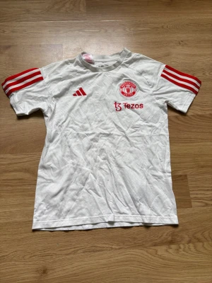 Manchester United vit träningströja - Säljer en vit Manchester United träningströja från Adidas med röda detaljer och tre ränder på ärmarna. Tröjan har klubbmärke, Adidas-logga och Tezos sponsortryck på bröstet. Tillverkad i mjukt och lätt material, perfekt för träning eller match.
