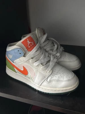 Nike Air Jordan 1 Mid multicolor - Nike Air Jordan 1 Mid sneakers i vitt läder med färgglada detaljer. Orange swoosh, blå och gröna paneler, broderade moln och gräsdetaljer. Klassisk siluett med perforerad tå och vit sula. Perfekt för dig som vill sticka ut med din stil.