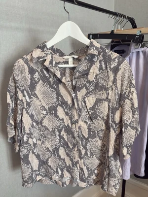 Beige och grå skjorta med ormmönster - Trendig skjorta från H&M med beige och grått ormmönster. Skjortan har korta ärmar, klassisk krage och knappar framtill. Perfekt för dig som vill sticka ut med ett djurmönster i garderoben.