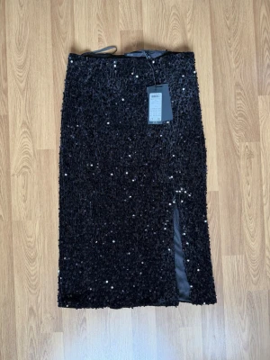 Svart paljettkjol med slits från Vero Moda - Säljer en svart långkjol från Vero Moda täckt med glittriga paljetter. Kjolen har en rak passform och en snygg slits framtill som ger extra edge. Perfekt för dig som vill sticka ut med en riktigt statement piece.