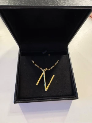 Maria Nilsdotter halsband  - Snyggt halsband i guldfärgad metall med ett stilrent hänge i form av bokstaven N. Kedjan är tunn och elegant, perfekt för att ge en personlig touch till din stil. Kommer i en svart smyckesask. Modellen wonky letter nacklace från Maria Nilsdotter. Halsbandet är i jättefint skick, boxen till halsbandet medföljer. Kan sänka priset något vid snabbt köp. Kedjan är 45cm. 