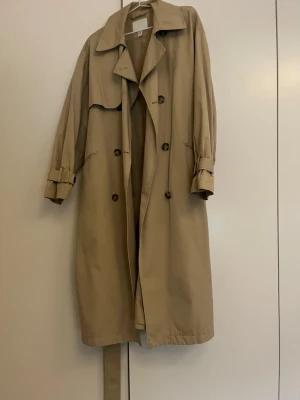 Beige klassisk trenchcoat - Stilren beige trenchcoat med dubbelknäppning, bred krage och bälte i midjan. Jackan har långa ärmar med justerbara spännen vid ärmsluten och typisk stormflik på ena sidan. Perfekt för dig som gillar tidlös och enkel stil.