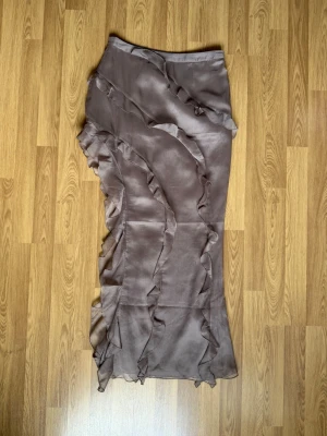 Brun volangkjol från H&M - Säljer en brun, lång kjol från H&M med snygga volangdetaljer längs sidorna och framtill. Kjolen har en lätt glansig finish och är tillverkad i ett tunt, flowy material som ger en cool och trendig look.