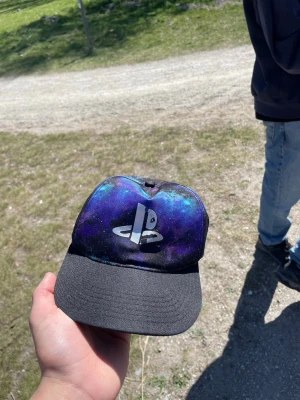 PlayStation keps med galaxmönster - Snygg keps från H&M med PlayStation-logga framtill och coolt galaxmönster i blått, lila och svart. Skärmen är svart och kepsen har justerbar rem bak. Perfekt för dig som gillar gaming och vill sticka ut med din stil.