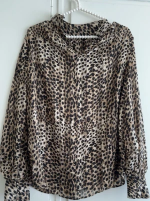 Leopardmönstrad blus med v-ringning - Snygg blus med leopardmönster i beige, brunt och svart. Blusen har v-ringning och långa ärmar med breda manschetter. Materialet är lätt och ger ett lyxigt intryck. Perfekt för dig som vill sticka ut med ett trendigt djurmönster.