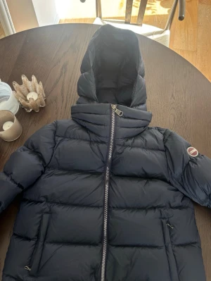 Mörkblå dunjacka från Colmar - Snygg och populär mörkblå dunjacka från Colmar med huva och dragkedja framtill. Jackan har två fickor med dragkedja och en rund logotyp på ärmen. Perfekt för kalla vinterdagar och riktigt skön att bära. Nypris 6000