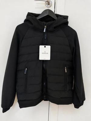 Svart pufferjacka från Moncler - Snygg svart pufferjacka från Moncler med huva och dragkedja framtill. Jackan har två fickor med dragkedja och Moncler-logga på ärmen. Perfekt för kalla dagar med sin vadderade design och stilrena look. Priset går att diskuteras vid en bra affär! 😊