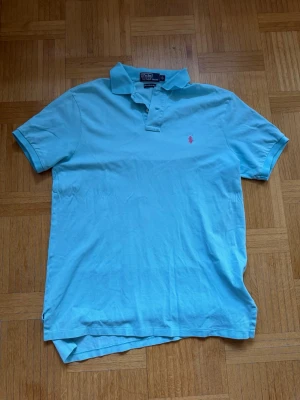 Ljusblå pikétröja från Ralph Lauren - Snygg ljusblå pikétröja från Ralph Lauren med klassisk krage och korta ärmar. Tröjan har en rosa broderad logga på bröstet och är tillverkad i mjuk bomull. Perfekt för en avslappnad och stilren look. Sparsamt använd, skriv vid funderingar ☺️