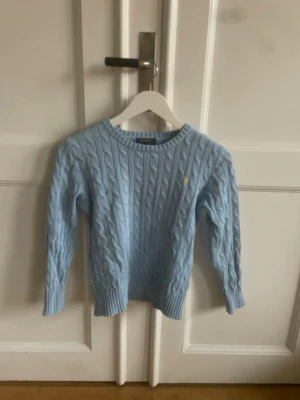 Ljusblå kabelstickad tröja Polo Ralph Lauren - Säljer en ljusblå kabelstickad tröja från Polo Ralph Lauren med rund halsringning och ribbade muddar. Tröjan har det klassiska broderade logot i gult på bröstet och är tillverkad i mjukt material, perfekt för lager på lager.