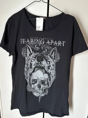 Svart t-shirt med vargtryck från H&M - Cool svart t-shirt från H&M Divided med stort grafiskt tryck av en varg och ormar samt texten 'After Darkness Fearing Apart' på bröstet. T-shirten har rund hals och korta ärmar. Perfekt för dig som gillar edgy och unika motiv.