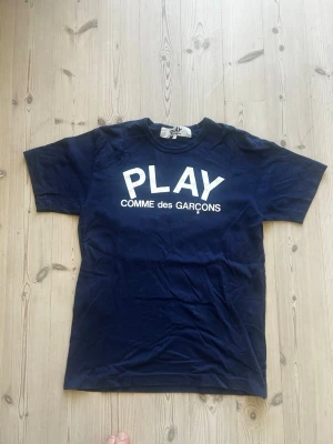 Blå Comme des Garçons PLAY t-shirt - Mörkblå t-shirt från Comme des Garçons PLAY med vit logotyptext framtill. Klassisk rund hals och korta ärmar. Tillverkad i japansk bomull. Storlek L men fits M då det är japansk storlek. Tröjan är som ny utan hål, fläckar eller tecken på användning.