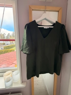 Mörkgrön v-ringad blus från Vero Moda - Snygg mörkgrön blus från vila med v-ringning och korta, vida ärmar. Tillverkad i ett lätt och mjukt material som ger ett elegant fall. Perfekt att styla med jeans eller kjol för en trendig look.