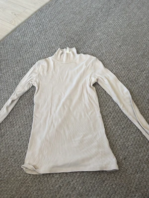 Beige ribbad polotröja - Säljer en beige ribbad polotröja med lång ärm och hög krage. Tröjan är figurnära och har en enkel, stilren design som passar perfekt till lager-på-lager. Materialet är mjukt och stretchigt, vilket gör den bekväm att bära.