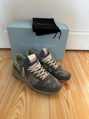 Philippe Model traniers  - Snygga sneakers från Philippe Model med camo-mönster i grått och grönt, ljusa snören och mintgrön detalj på hälen. Skorna har en låg profil och är tillverkade i en mix av mocka och textil. Perfekta för dig som vill ha en unik och trendig look.