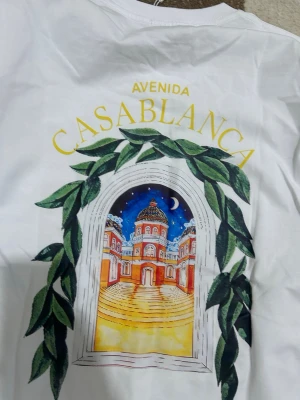 Vit Avenida Casablanca t-shirt - Vit t-shirt från Casablanca med färgglad grafisk print på ryggen som föreställer en arkitektonisk scen omgiven av gröna blad och texten 'Avenida Casablanca' i gult. Klassisk passform och korta ärmar, perfekt för dig som gillar unika prints. Kan bytas mot burberry t shirt
