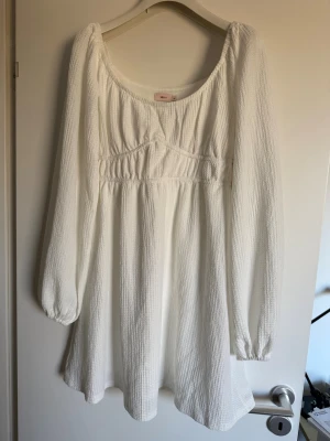 Vit kortklänning med lång puffärm - Supersöt vit kortklänning från Nelly med långärmade puffärmar och en lätt strukturerad yta. Klänningen har en markerad midja och rundad urringning, perfekt för sommarens alla tillfällen. Tillverkad i ett mjukt och luftigt material i polyester/ elastan. Oanvänd 