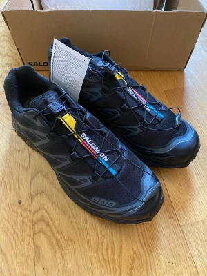 Salomon svart S/LAB sneakers - Svarta Salomon S/LAB sneakers med mesh- och syntetmaterial, snabbsnörning och färgdetaljer i gult, rött och blått på plösen. Skorna har en sportig design med robust sula och diskreta grå loggor på sidan. Perfekta för dig som gillar streetwear och outdoor-vibe.