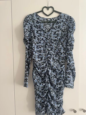Blommig blå långärmad klänning - Snygg kortklänning med blått och svart blommönster. Klänningen har långa ärmar, rynkade detaljer framtill och en figurnära passform. Perfekt för dig som gillar en feminin och trendig stil.