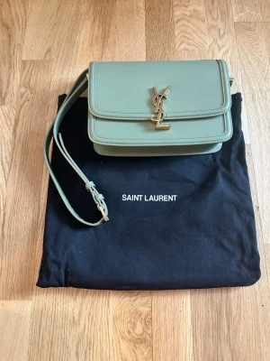 Grön axelväska från Saint Laurent - Snygg grön axelväska från Saint Laurent i slätt läder med justerbar axelrem och ikoniskt YSL-spänne i guld framtill. Väskan har en stilren, rektangulär form och levereras med original dustbag. Perfekt för dig som gillar lyxiga accessoarer.