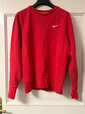 Röd stickad tröja från Nike - Röd stickad tröja från Nike med rund halsringning och vit broderad logga på bröstet. Tröjan är tillverkad i 100% bomull och har ribbade muddar vid ärmslut och nederkant. Perfekt för en avslappnad och sportig stil.