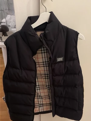 Burberry väst - Pris går absolut att diskuteras och billigare vid snabb affär.Snygg svart dunväst från Burberry med klassiskt rutigt foder på insidan och diskret logga på bröstet. Västen har hög krage och quiltad design som ger en modern look. Perfekt för lager-på-lager och enkel att matcha med olika outfits.