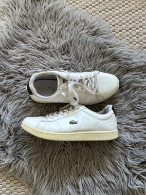 Vita Lacoste sneakers med logga - Klassiska vita sneakers från Lacoste med den ikoniska gröna krokodilloggan på sidan. Skorna har lågt skaft, vita snören och diskreta sömmar. Ovandelen är i slätt läder och sulan är vit med en lätt gulnad nyans.